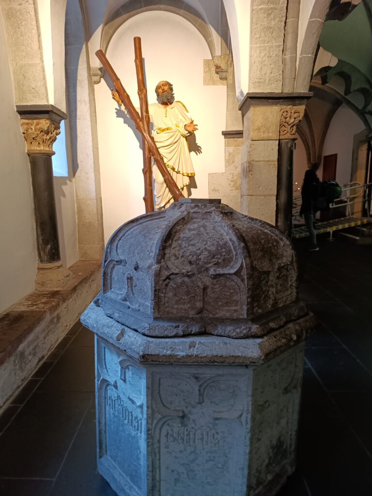 Der Blutbrunnen in der Kirche Sankt Andreas, Köln, im Kontext der Kirchenarchitektur mit einer Heiligenfigur im Hintergrund.